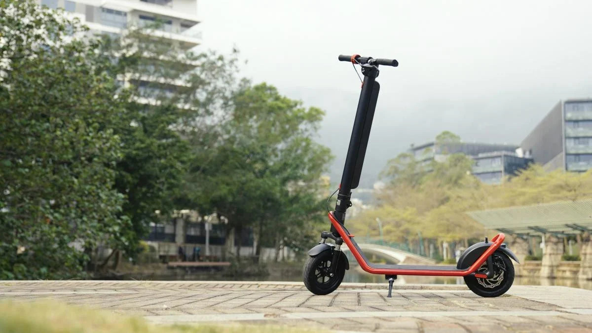 Black HX electric scooter with a red frame on its kickstand on a paved promenade, with trees and modern buildings behind