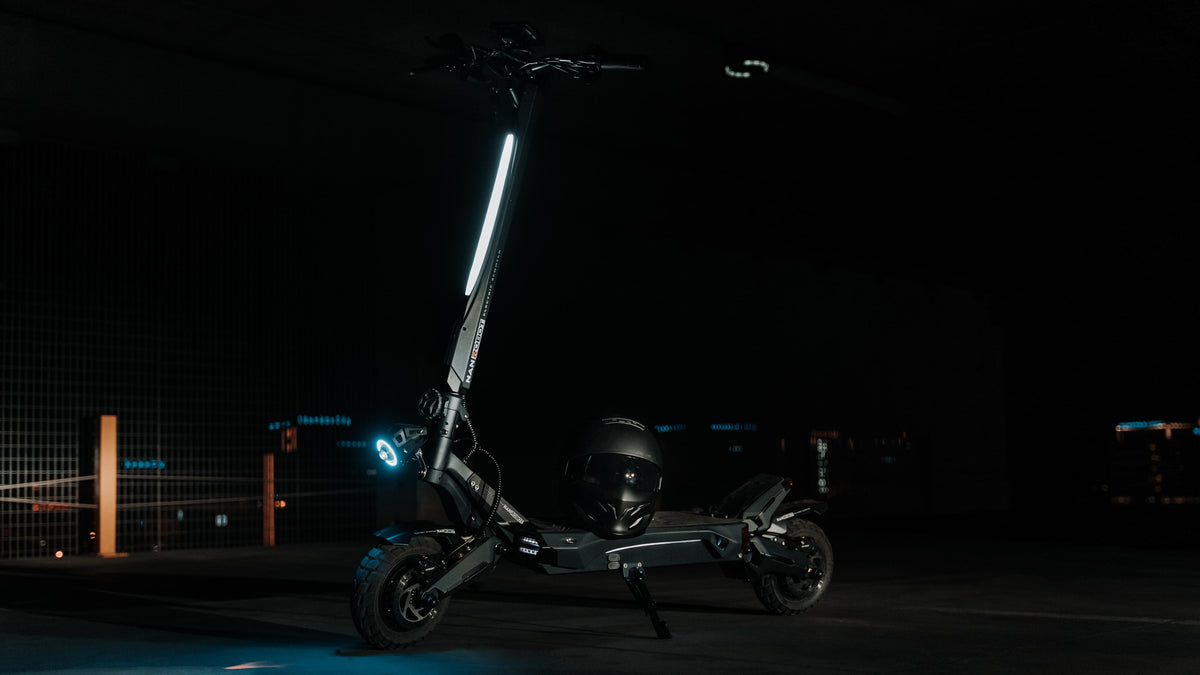 Black electric scooter parked on its kickstand in a dark garage, with a helmet resting on the deck