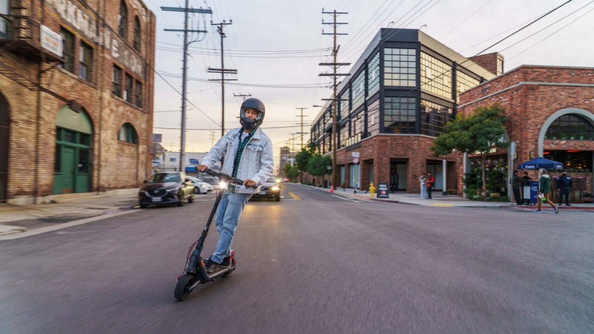Man in a black full-face helmet riding an electric scooter with its headlight on down a city street at dusk