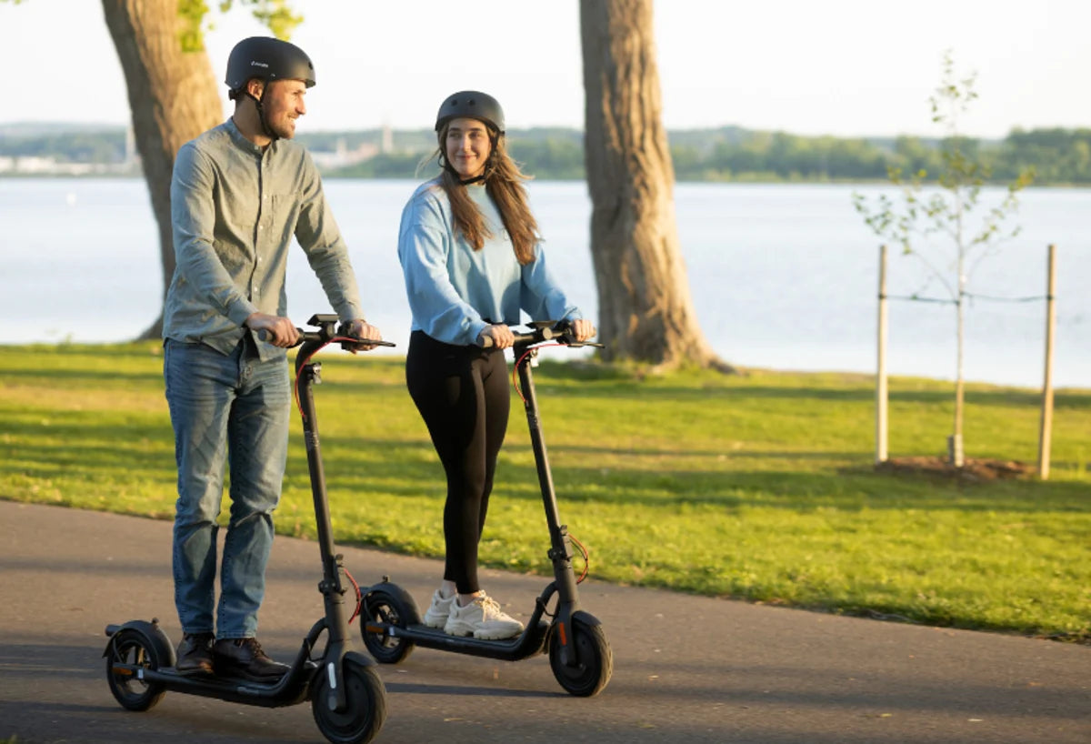 Two riders wearing helmets on NAVEE electric scooters on a paved path beside a lake