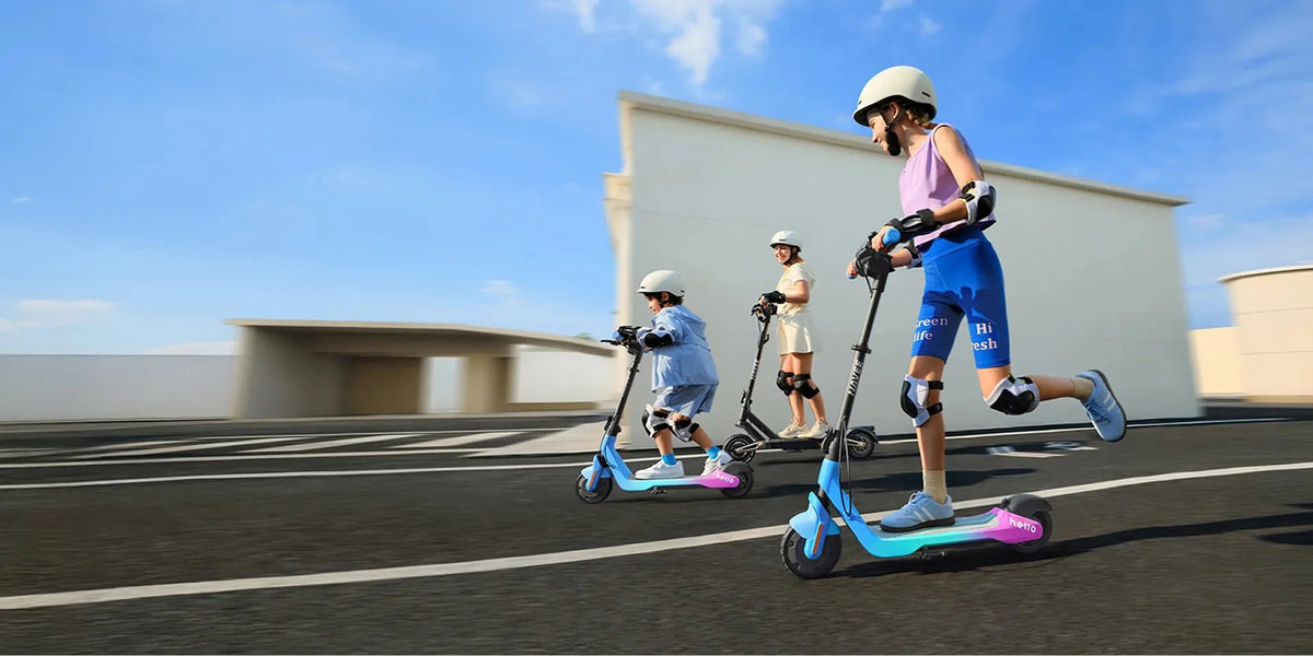 Three children wearing helmets, knee and elbow pads ride electric scooters across a sunny asphalt square
