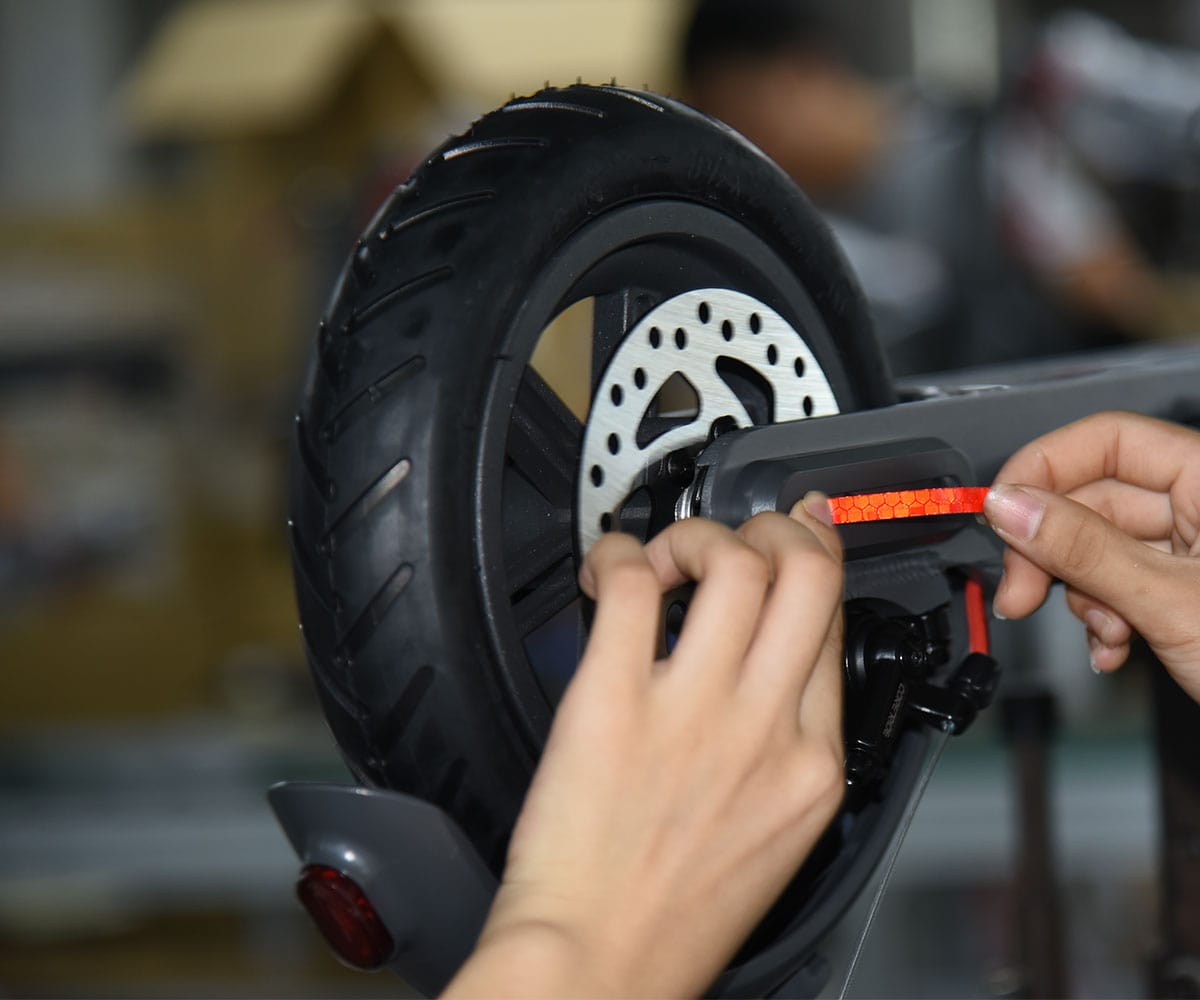 Hands fitting a red reflective strip to the rear fork of an electric scooter, beside the treaded tyre and disc brake