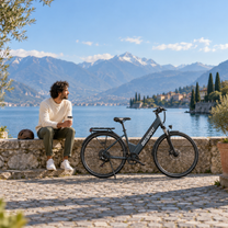 OneSport OT12 Pro electric bike on a paved lakeside quay, with a man sitting on the wall beside it and mountains in the background
