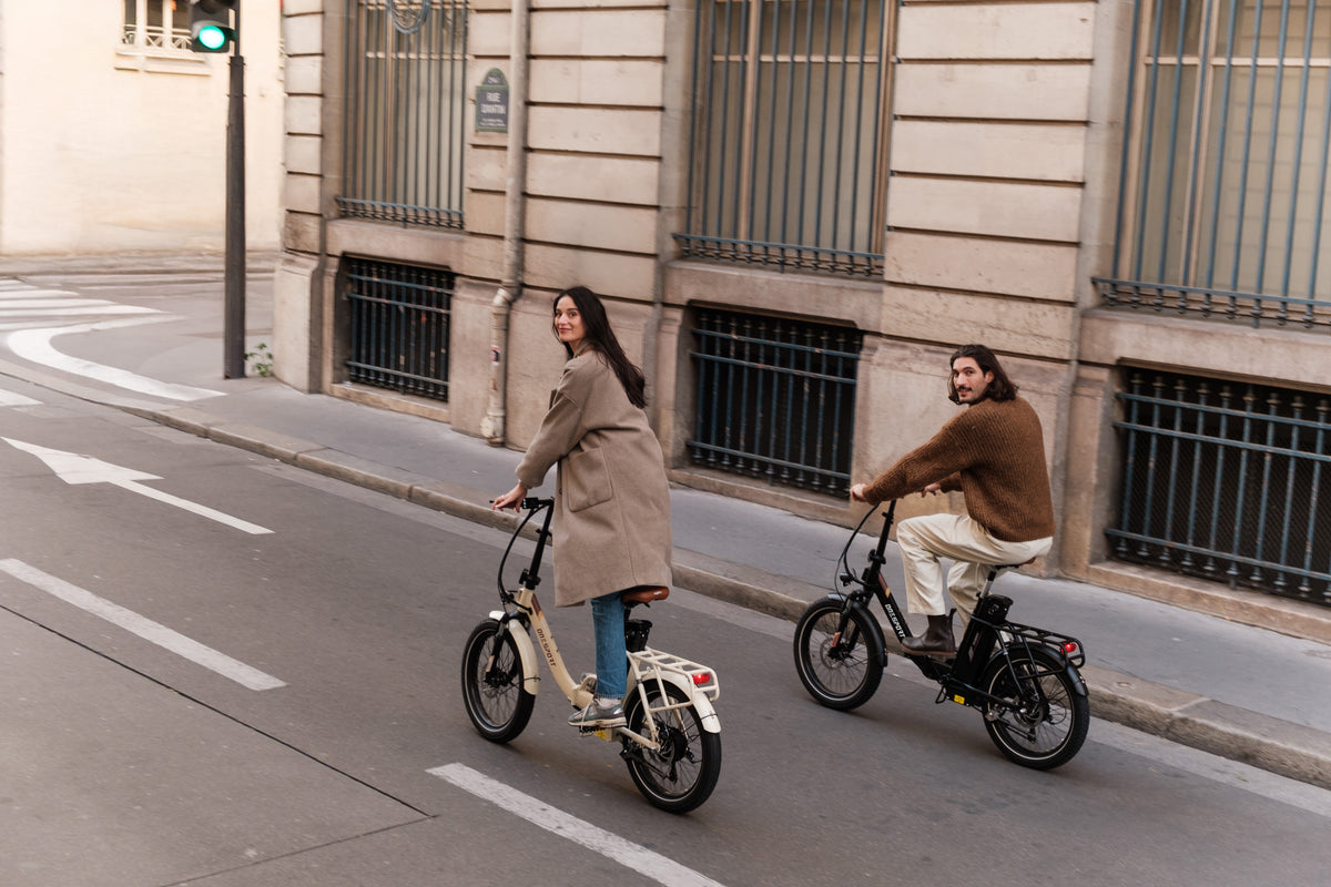 Two people riding folding bikes on a city street, one beige and one black