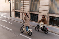Two people riding folding bikes on a city street, one beige and one black