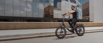 Man in sunglasses riding the OneSport OT16 Pro along a street in front of a glass facade