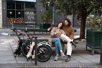 A couple on a bench next to two folded OneSport bikes, one beige and one black
