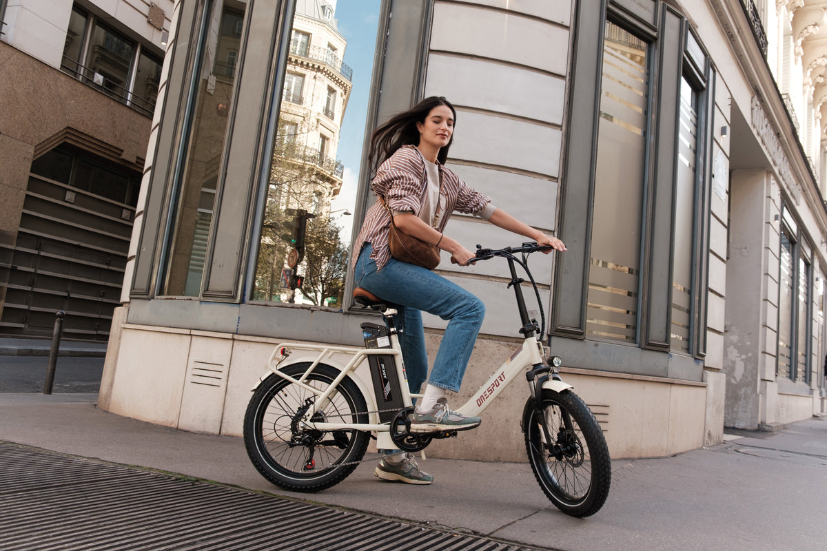 Woman riding the OneSport OT16 Pro, a white electric bike, side view on a city street by a stone building