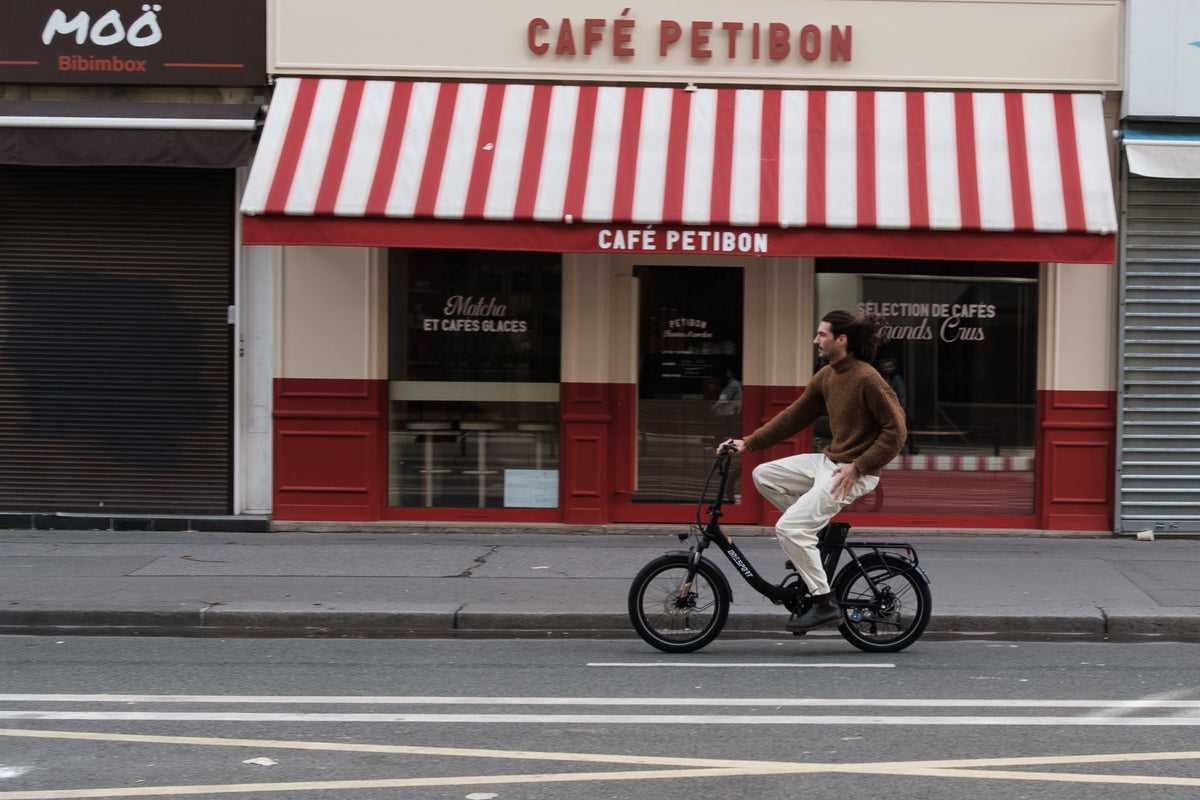 Man riding the OneSport OT16 Pro, a black bike, in side view in front of a cafe with a red-and-white striped awning
