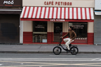 Man riding the OneSport OT16 Pro, a black bike, in side view in front of a cafe with a red-and-white striped awning