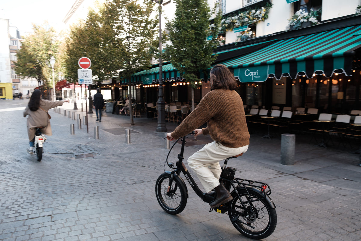 Person riding the OneSport OT16 Pro, a black bike, on a cobblestone street past a cafe with a green-striped awning
