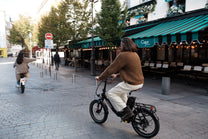 Person riding the OneSport OT16 Pro, a black bike, on a cobblestone street past a cafe with a green-striped awning