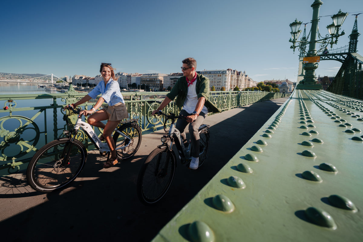 Two people cycling on a green bridge in sunshine with buildings and water in the background