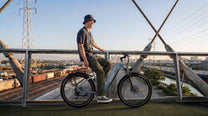 Smiling man in a hat sitting on OneSport WT05 by a bridge railing with railway tracks and a city in the background