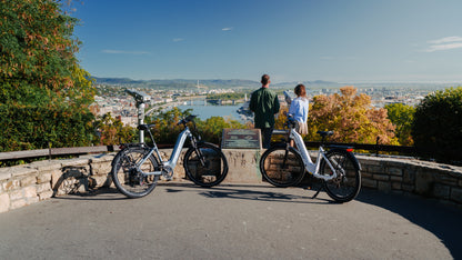 Two OneSport WT05 bikes parked by a stone railing at a viewpoint with two people and a view over the city and river