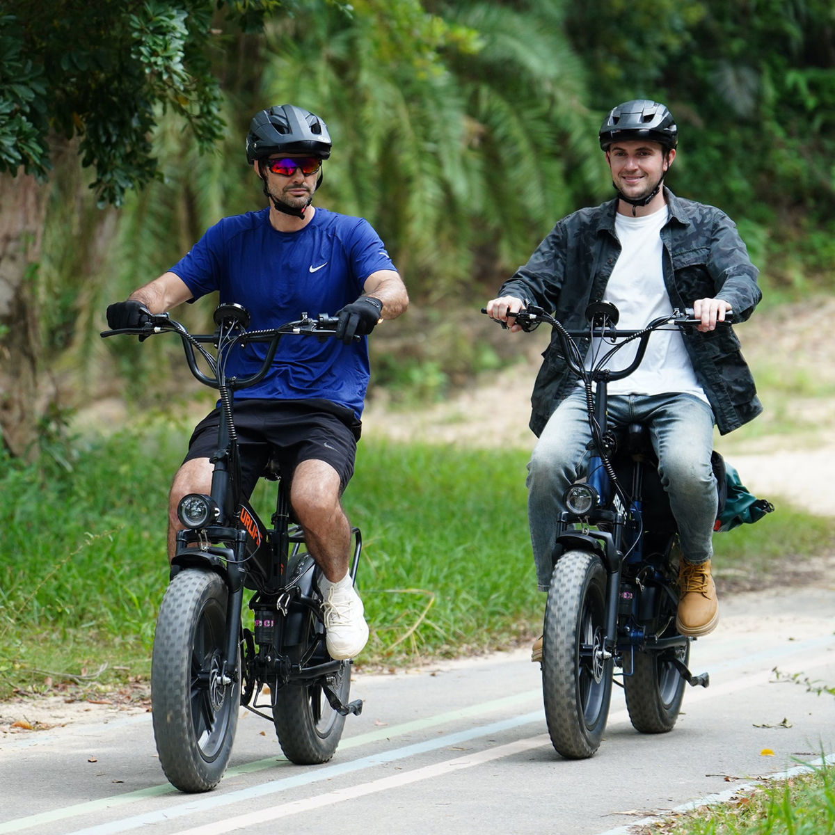 Two people in helmets riding side by side on fat-tyre bikes on a tarmac road surrounded by greenery