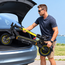 Man wearing sunglasses lifting a folded black and yellow electric scooter with the text FREEBOY at the open trunk of a gray car