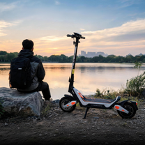 Wheely - J18 on its kickstand by a lake at sunset, with a person wearing a backpack sitting on a rock nearby
